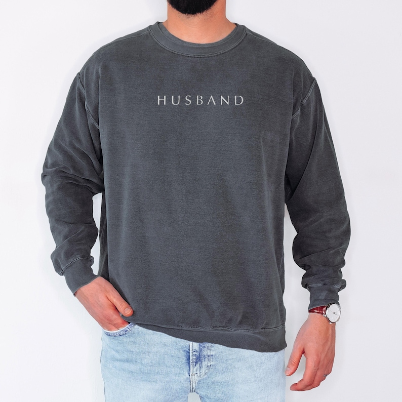 May include: A dark grey sweatshirt with a white embroidered word "HUSBAND" on the front. The sweatshirt has a relaxed fit and long sleeves.