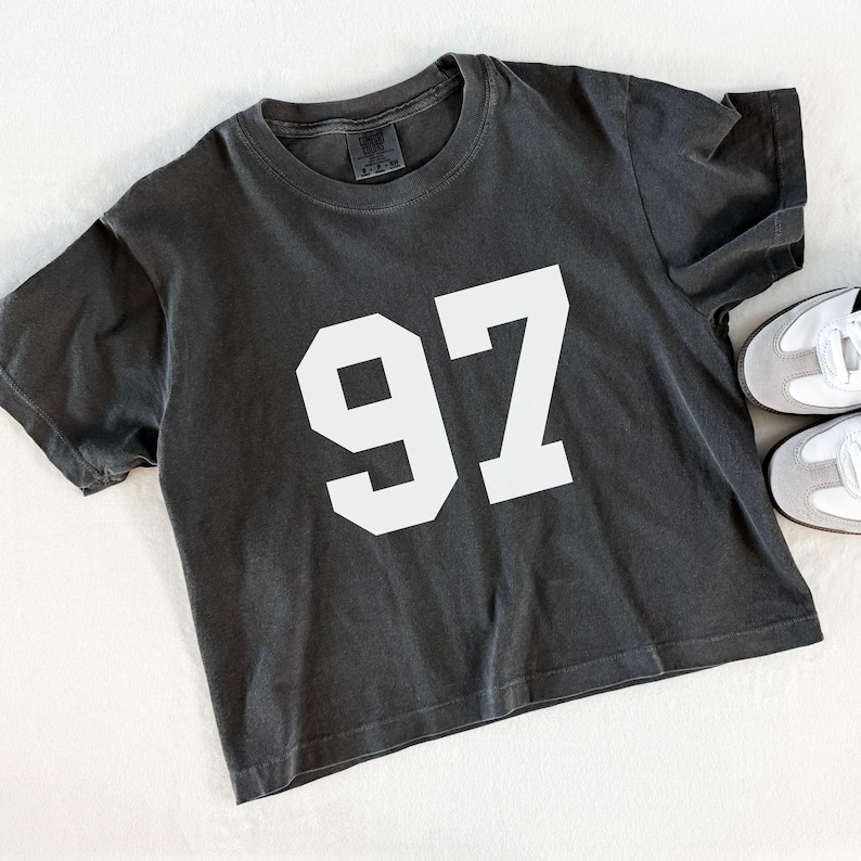 May include: A charcoal gray t-shirt with the number "97" in white block letters. The shirt has short sleeves and a crew neck. A pair of white and gray sneakers are next to the shirt.
