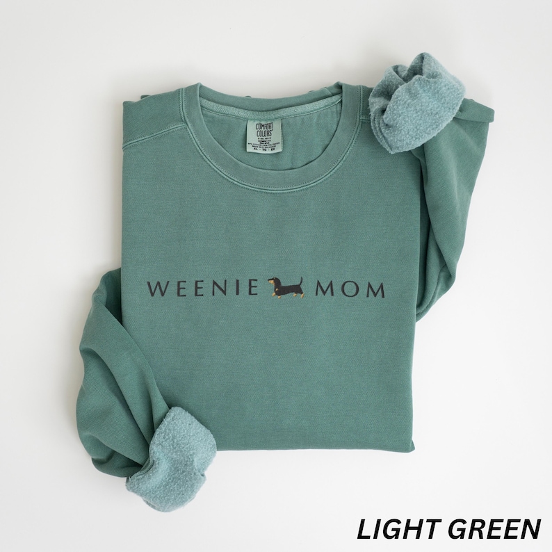 Puede incluir: Sudadera verde claro con el texto "WEENIE MOM" y una ilustraci&oacute;n de perro salchicha. La sudadera tiene cuello redondo y mangas largas. La parte inferior de la imagen tiene el texto "LIGHT GREEN".