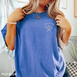 May include: A blue t-shirt with the text "CUSTOM" above a tennis ball and crossed rackets design. The shirt also has the text "FLO BLUE" at the bottom. The shirt is a solid blue color.