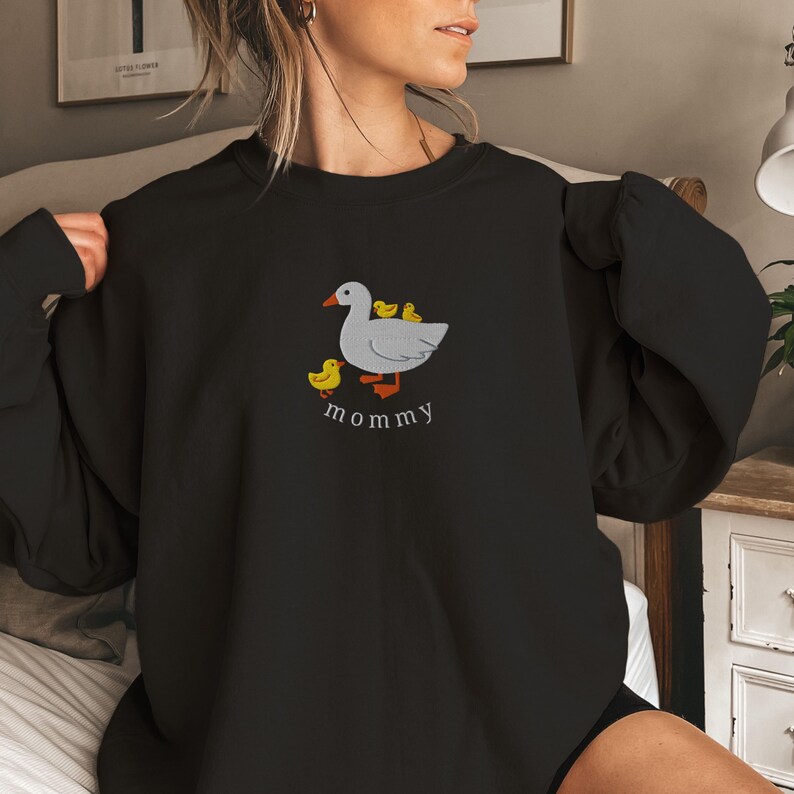 May include: Black sweatshirt with a white goose and three yellow ducklings embroidered on the front. The text "mommy" is embroidered below the goose.