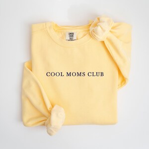 May include: A pale yellow sweatshirt with the navy blue text "COOL MOMS CLUB". The sweatshirt is folded with a knot tied in the sleeve. The Comfort Colors brand label is visible at the neckline.