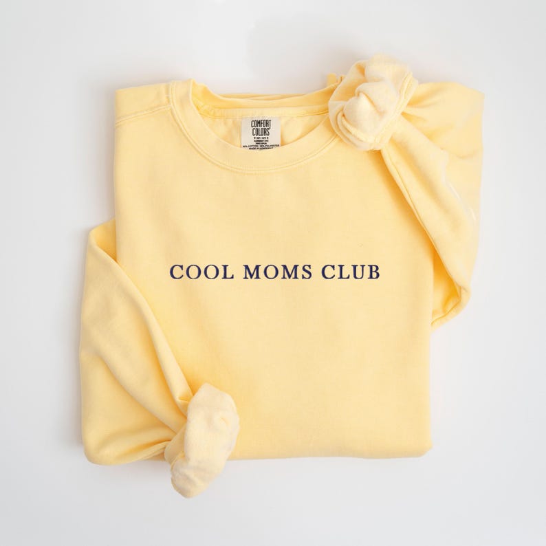 May include: A pale yellow sweatshirt with the navy blue text "COOL MOMS CLUB". The sweatshirt is folded with a knot tied in the sleeve. The Comfort Colors brand label is visible at the neckline.