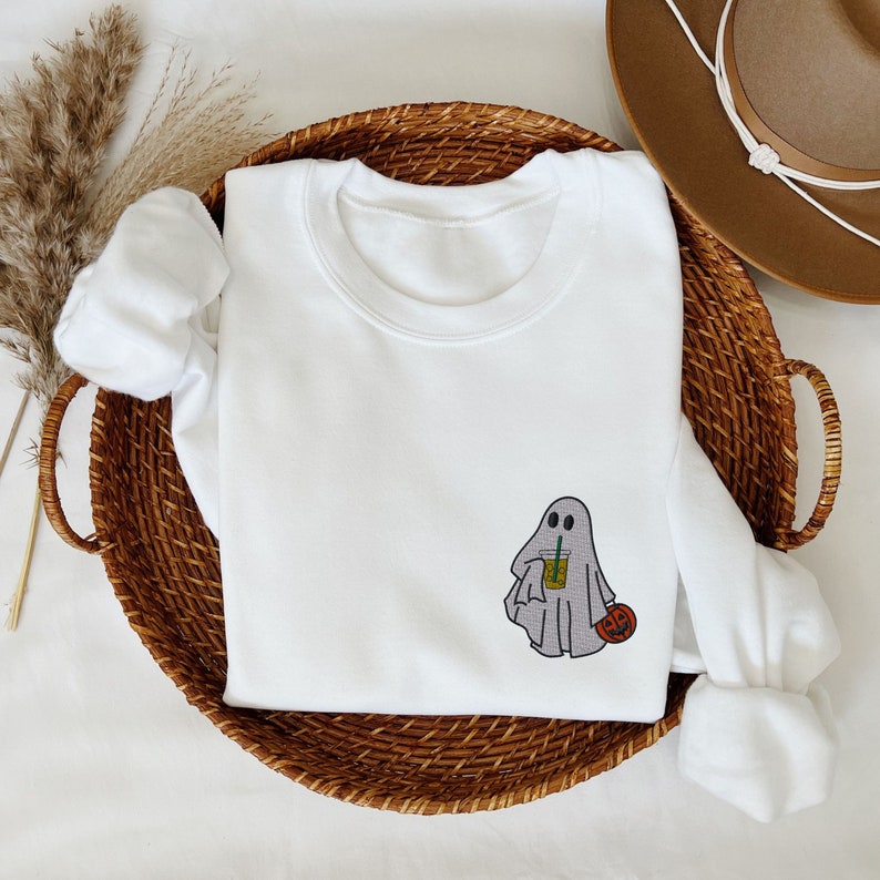 May include: White sweatshirt with a ghost holding a beverage and a small pumpkin. The ghost is gray and has two eyes. The pumpkin is orange.
