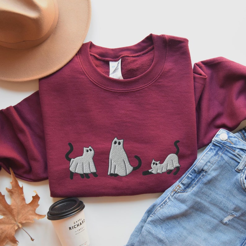 K&ouml;nnte beinhalten: Ein weinroter Pullover mit einem Aufdruck von drei Katzen, die als Geister verkleidet sind. Die Katzen sind wei&szlig; mit schwarzen Umrissen und in verschiedenen Posen. Der Pullover liegt auf einer wei&szlig;en Oberfl&auml;che mit einem braunen Hut, einer blauen Jeans und einer Kaffeetasse.