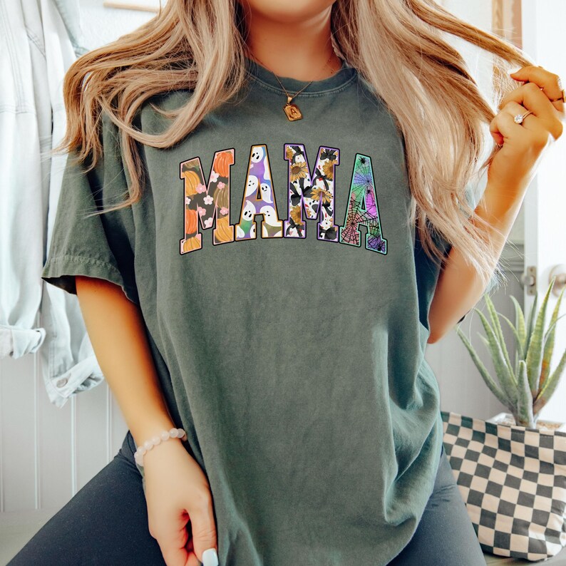 Puede incluir: Una camiseta verde oscuro con la palabra "MAMA" impresa en letras grandes y coloridas. Las letras est&aacute;n decoradas con una variedad de patrones de tem&aacute;tica de Halloween, incluyendo calabazas, fantasmas, murci&eacute;lagos y ara&ntilde;as.
