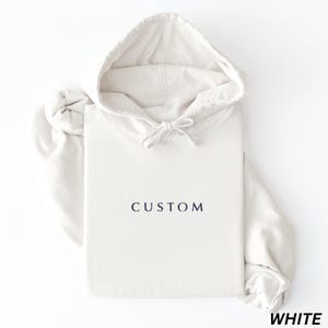 May include: A white hoodie with the word "CUSTOM" printed in navy blue. The hoodie has a drawstring hood and long sleeves. The word "WHITE" is printed in the bottom right corner.