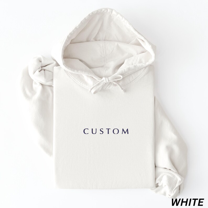 May include: A white hoodie with the word "CUSTOM" printed in navy blue. The hoodie has a drawstring hood and long sleeves. The word "WHITE" is printed in the bottom right corner.