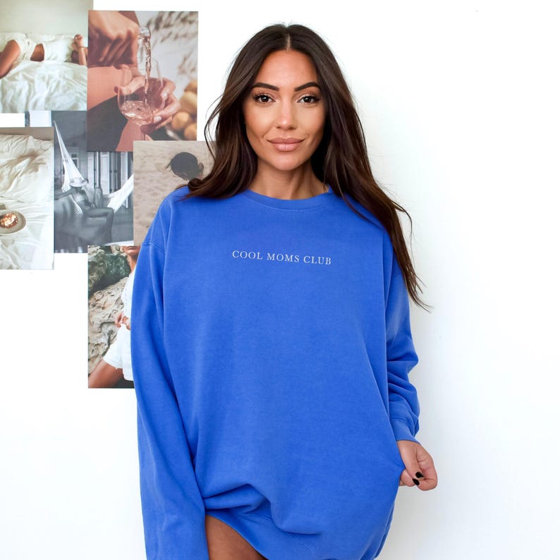 May include: A woman wearing a blue sweatshirt with the text "COOL MOMS CLUB" in white. The sweatshirt is a solid, vibrant blue colour. The woman has long, dark hair and is smiling. A collage of images is in the background.