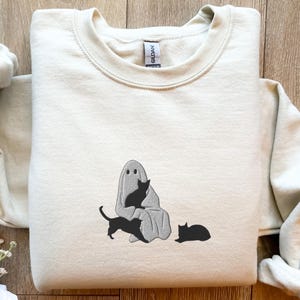 K&ouml;nnte beinhalten: Ein hellbeiger Pullover mit einem schwarzen und wei&szlig;en gestickten Design eines Geistes mit drei schwarzen Katzen. Der Geist tr&auml;gt ein Laken und hat zwei Augen. Eine Katze sitzt auf dem Scho&szlig; des Geistes, eine andere Katze sitzt vor dem Geist und die dritte Katze liegt auf dem Boden.