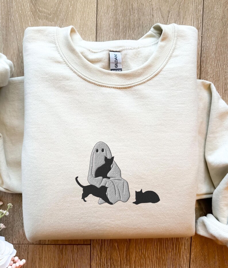 K&ouml;nnte beinhalten: Ein hellbeiger Pullover mit einem schwarzen und wei&szlig;en gestickten Design eines Geistes mit drei schwarzen Katzen. Der Geist tr&auml;gt ein Laken und hat zwei Augen. Eine Katze sitzt auf dem Scho&szlig; des Geistes, eine andere Katze sitzt vor dem Geist und die dritte Katze liegt auf dem Boden.
