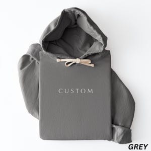 May include: A gray hoodie with a drawstring hood and the word "CUSTOM" embroidered on the front. The sleeves are cuffed, and the hoodie is folded. The word "GREY" is printed in the bottom right corner.