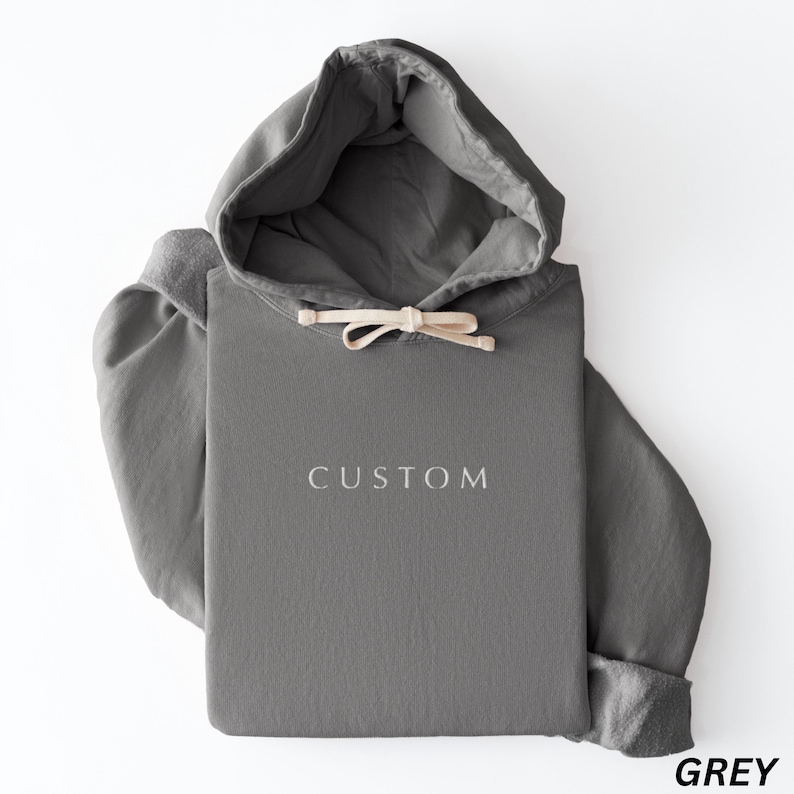 May include: A gray hoodie with a drawstring hood and the word "CUSTOM" embroidered on the front. The sleeves are cuffed, and the hoodie is folded. The word "GREY" is printed in the bottom right corner.