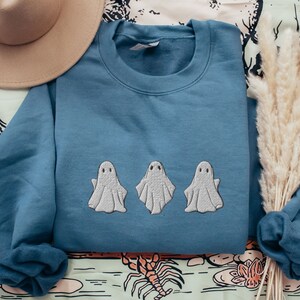 May include: A blue sweatshirt with three white embroidered ghosts on the front. The ghosts are in a row, facing forward, with their arms outstretched.