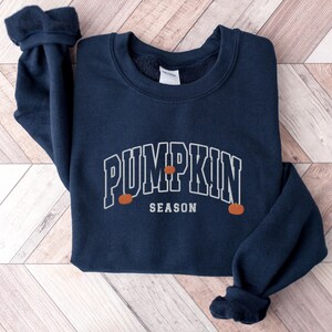 May include: A navy blue sweatshirt with the text "Pumpkin Season" in white outlined letters. Three small orange pumpkins are depicted on the sweatshirt.