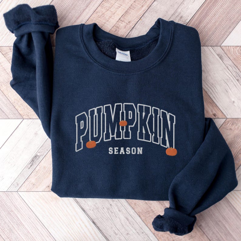 May include: A navy blue sweatshirt with the text "Pumpkin Season" in white outlined letters. Three small orange pumpkins are depicted on the sweatshirt.