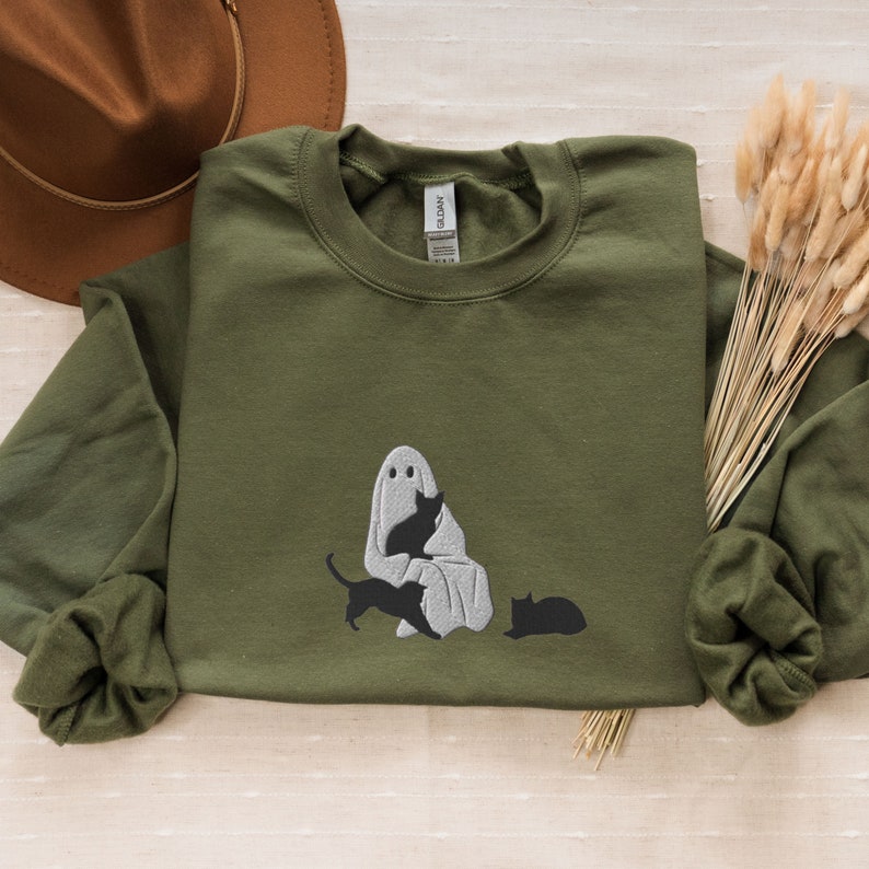 K&ouml;nnte beinhalten: Ein gr&uuml;nes Sweatshirt mit einem gestickten Design eines Geistes, der eine schwarze Katze h&auml;lt. Zwei weitere schwarze Katzen sitzen auf dem Boden unter dem Geist.