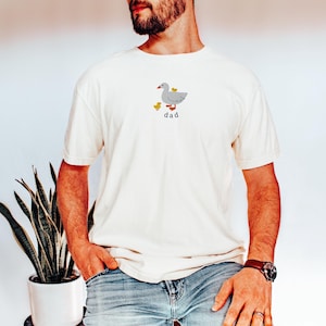 May include: Cream-colored t-shirt featuring a gray goose with two yellow ducklings and the word "dad" in black. The shirt is made of a soft material and has short sleeves.