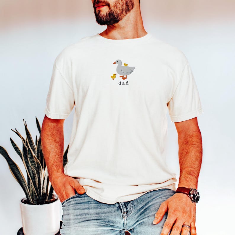 May include: Cream-colored t-shirt featuring a gray goose with two yellow ducklings and the word "dad" in black. The shirt is made of a soft material and has short sleeves.