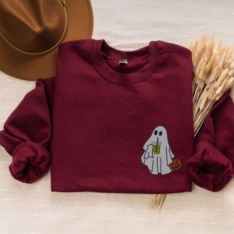 May include: A maroon sweatshirt with a white ghost holding a pumpkin and a beverage. The ghost has two eyes and a small mouth.