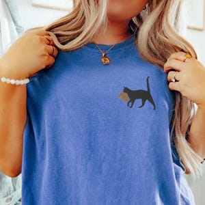 May include: A blue t-shirt with a black cat carrying a brown object in its mouth. The cat is on the left side of the shirt.