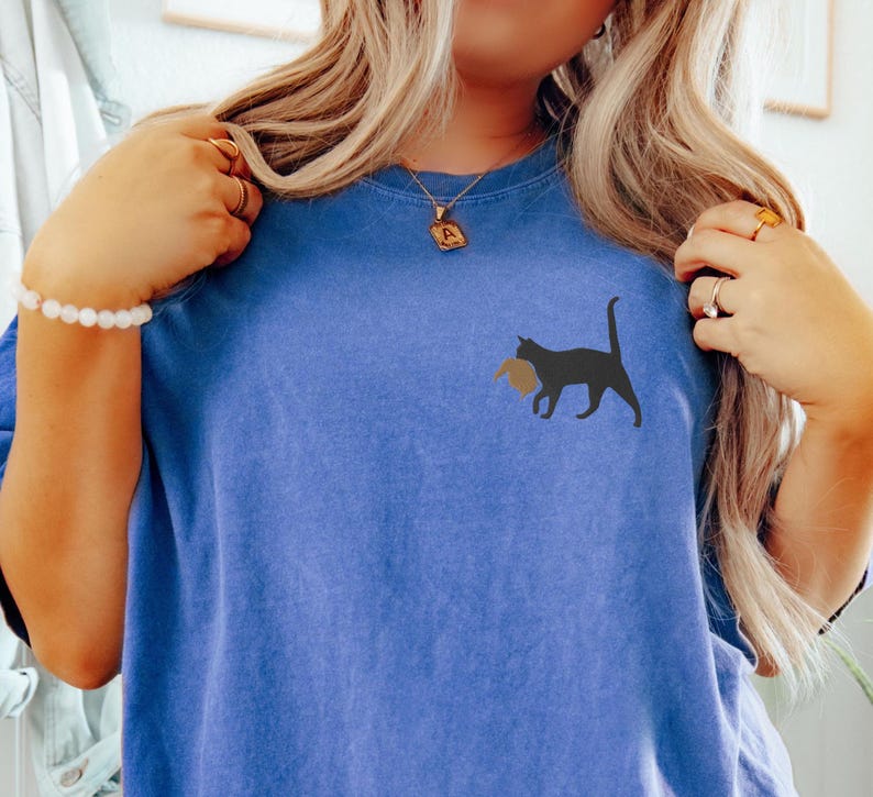May include: A blue t-shirt with a black cat carrying a brown object in its mouth. The cat is on the left side of the shirt.