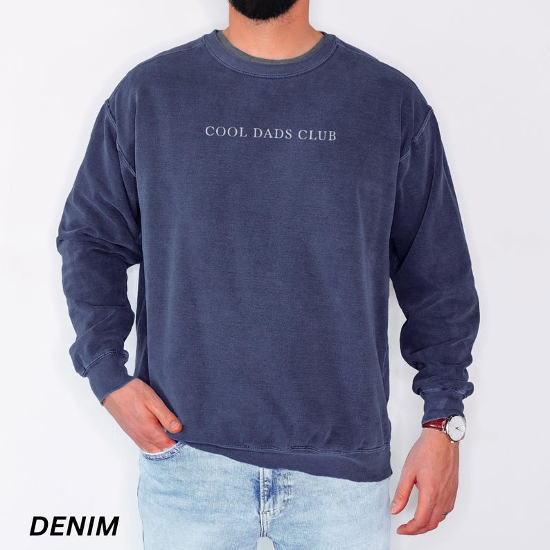 May include: A denim blue sweatshirt with the text "COOL DADS CLUB" in white. The sweatshirt has a crew neck and long sleeves. The word "DENIM" is printed at the bottom of the image.