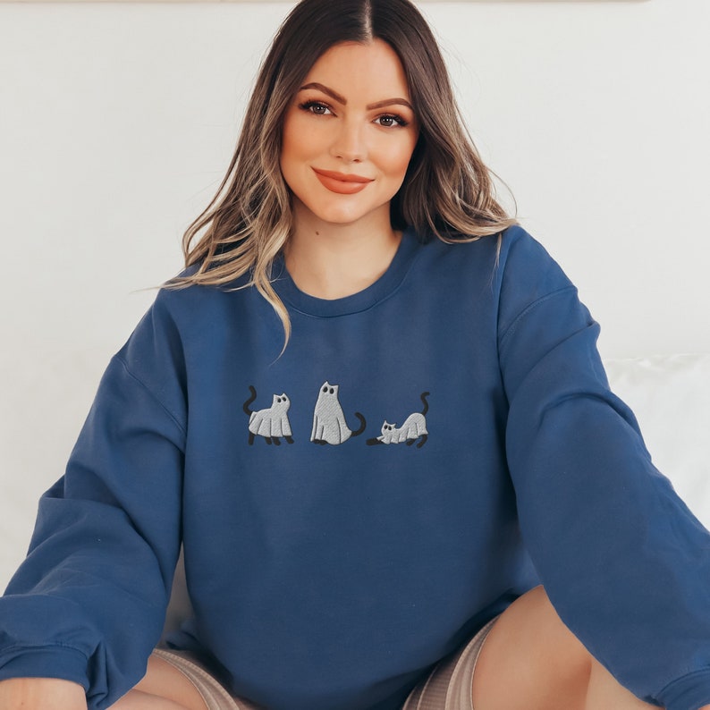 K&ouml;nnte beinhalten: Ein marineblauer Pullover mit einem Design aus drei grauen Katzen in einer Reihe. Die Katzen tragen Geisterkost&uuml;me f&uuml;r Halloween.
