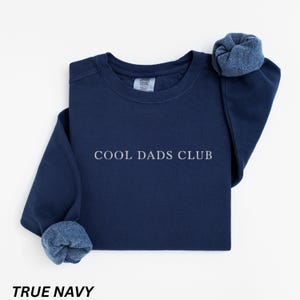 May include: A navy blue sweatshirt with the text "COOL DADS CLUB" in white. The sleeves are cuffed, revealing a lighter blue interior. The words "TRUE NAVY" are printed at the bottom of the image.