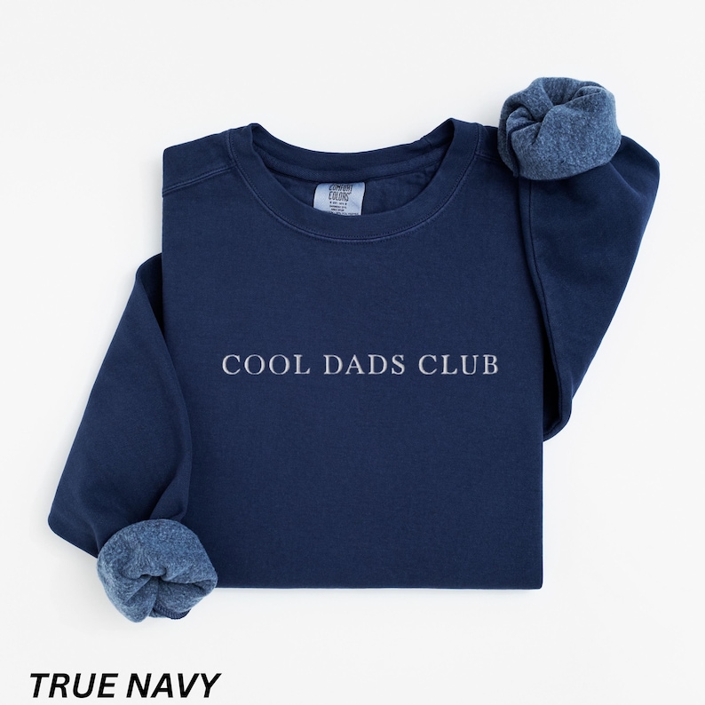 May include: A navy blue sweatshirt with the text "COOL DADS CLUB" in white. The sleeves are cuffed, revealing a lighter blue interior. The words "TRUE NAVY" are printed at the bottom of the image.