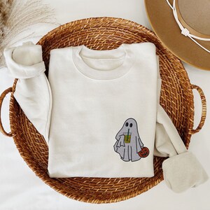 May include: A white sweatshirt with a ghost holding a cup of iced coffee and a small pumpkin. The ghost has two eyes and a small smile.