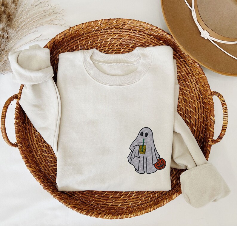 May include: A white sweatshirt with a ghost holding a cup of iced coffee and a small pumpkin. The ghost has two eyes and a small smile.