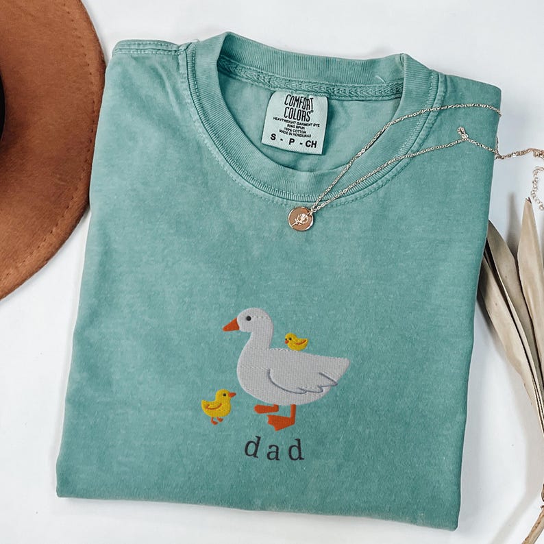 May include: A seafoam green Comfort Colors t-shirt featuring an embroidered design of a white duck with two yellow ducklings and the word "dad" in a simple font. A silver necklace with a pendant is draped across the shirt.
