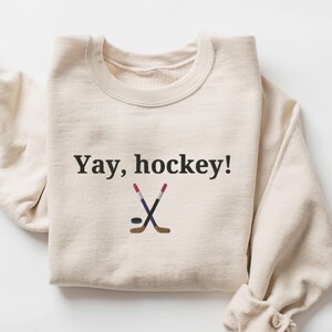 Puede incluir: Una sudadera de color crema con el texto "Yay, hockey!" y un gr&aacute;fico de dos palos de hockey cruzados y un disco.
