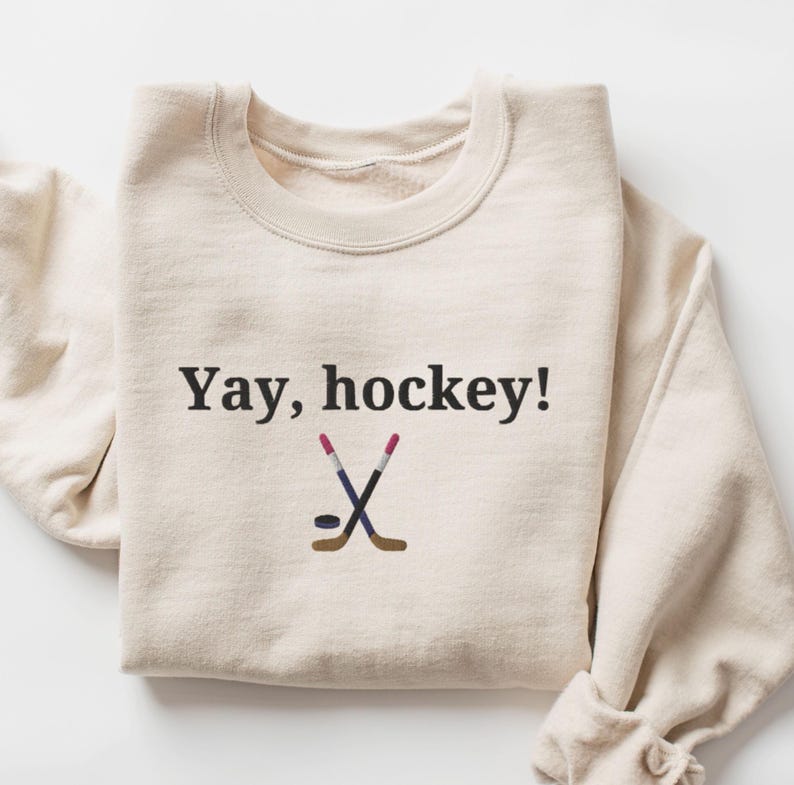 Puede incluir: Una sudadera de color crema con el texto "Yay, hockey!" y un gr&aacute;fico de dos palos de hockey cruzados y un disco.
