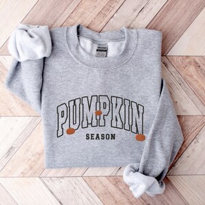 May include: A gray sweatshirt with the text "Pumpkin Season" in a black, collegiate-style font. Three small orange pumpkins are depicted on either side of the text.