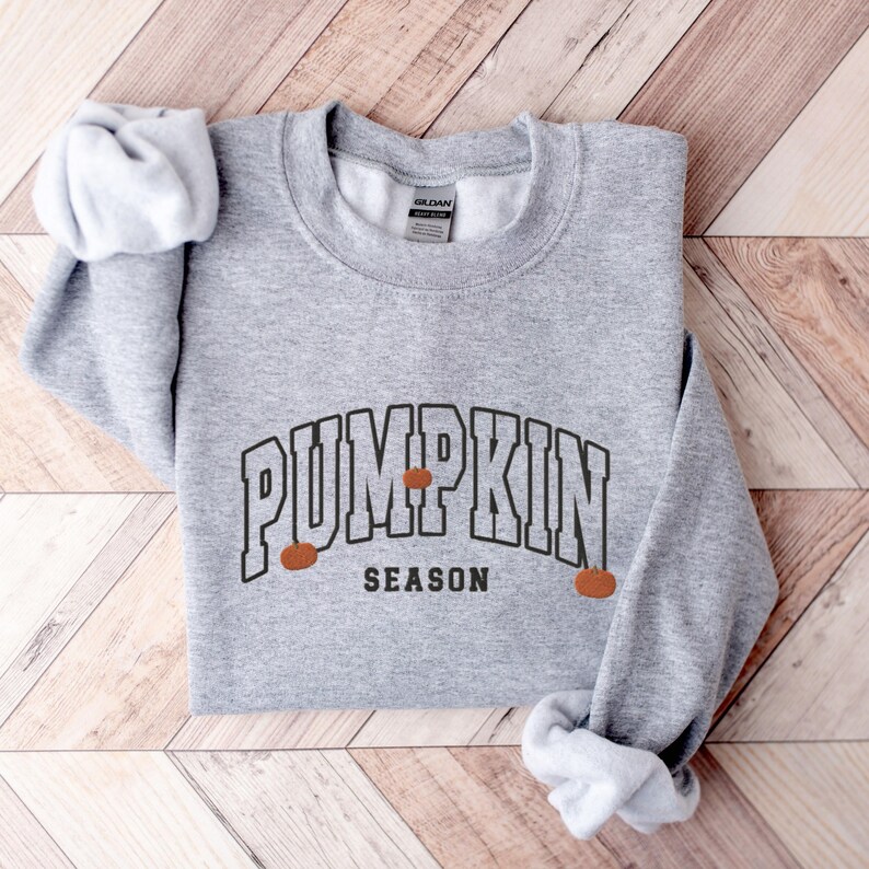 May include: A gray sweatshirt with the text "Pumpkin Season" in a black, collegiate-style font. Three small orange pumpkins are depicted on either side of the text.