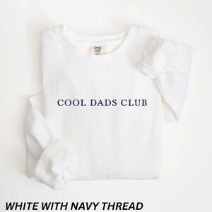 May include: A white sweatshirt with the text "COOL DADS CLUB" in navy blue. The sweatshirt has a crew neck and long sleeves. The text "WHITE WITH NAVY THREAD" is at the bottom of the image.
