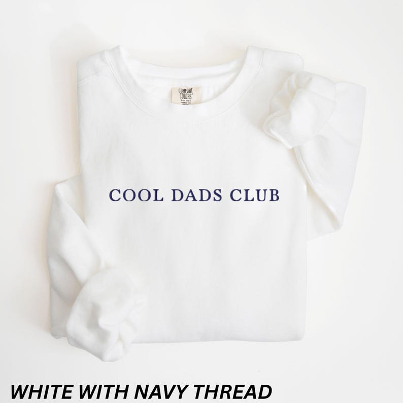 May include: A white sweatshirt with the text "COOL DADS CLUB" in navy blue. The sweatshirt has a crew neck and long sleeves. The text "WHITE WITH NAVY THREAD" is at the bottom of the image.