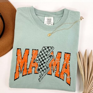 K&ouml;nnte beinhalten: Ein hellblaues T-Shirt mit einer abgenutzten orangefarbenen Grafik, auf der "MAMA" mit einem karierten Blitz in der Mitte steht. Das Shirt hat einen Rundhalsausschnitt und kurze &Auml;rmel.