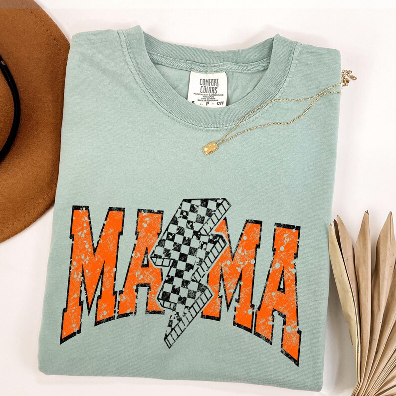 K&ouml;nnte beinhalten: Ein hellblaues T-Shirt mit einer abgenutzten orangefarbenen Grafik, auf der "MAMA" mit einem karierten Blitz in der Mitte steht. Das Shirt hat einen Rundhalsausschnitt und kurze &Auml;rmel.