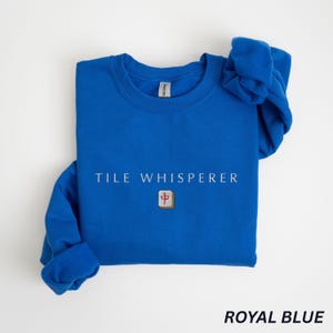 May include: Royal blue crewneck sweatshirt with the text "TILE WHISPERER" in white. A small, square tile with a red and brown design is centered below the text. The words "ROYAL BLUE" are printed in a sans-serif font at the bottom.