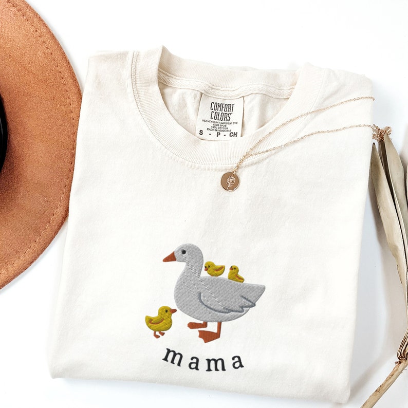 May include: A cream colored t-shirt with an embroidered design of a gray goose with three yellow ducklings. The text "mama" is embroidered below the goose.