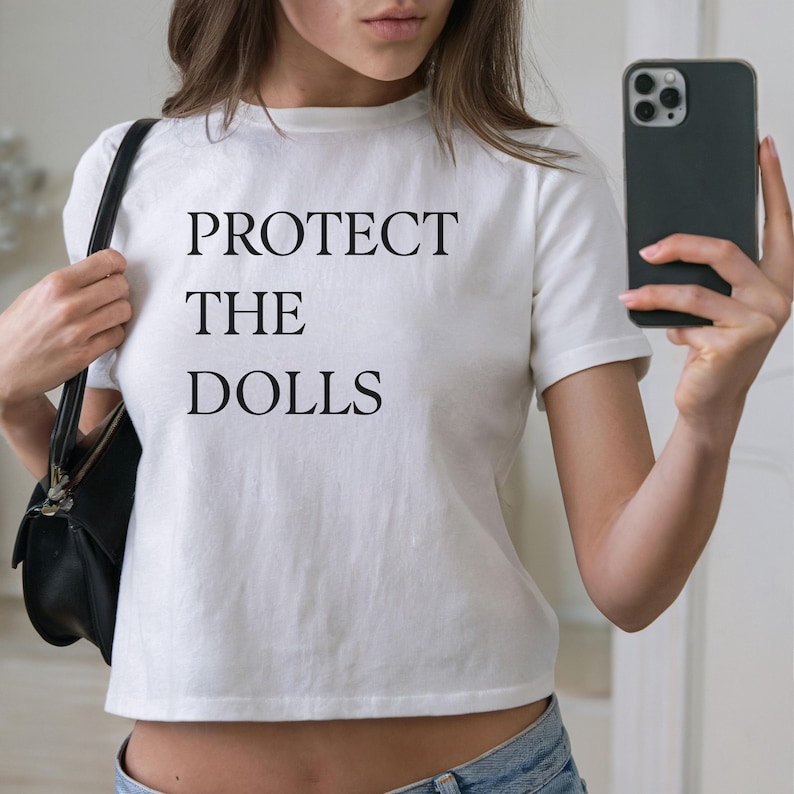 May include: White crop top with the text "PROTECT THE DOLLS" in black. The shirt is short-sleeved and has a crew neck. A black purse is on the left shoulder. A person is holding a black phone in the right hand.