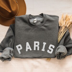 Puede incluir: Una sudadera gris oscuro con la palabra "PARIS" impresa en blanco en la parte delantera. La sudadera est&aacute; enrollada en las mangas y est&aacute; sobre una superficie beige claro.