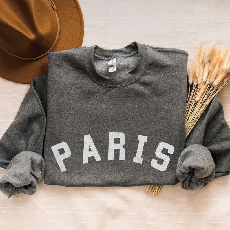 Puede incluir: Una sudadera gris oscuro con la palabra "PARIS" impresa en blanco en la parte delantera. La sudadera est&aacute; enrollada en las mangas y est&aacute; sobre una superficie beige claro.