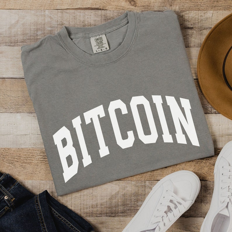 May include: A gray t-shirt with the word "BITCOIN" printed in white in a collegiate-style font.