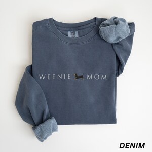 Puede incluir: Una sudadera azul denim con las palabras "WEENIE MOM" en letras blancas y una peque&ntilde;a ilustraci&oacute;n de perro salchicha. La sudadera tiene cuello redondo y mangas largas con pu&ntilde;os. La palabra "DENIM" est&aacute; impresa en la parte inferior.