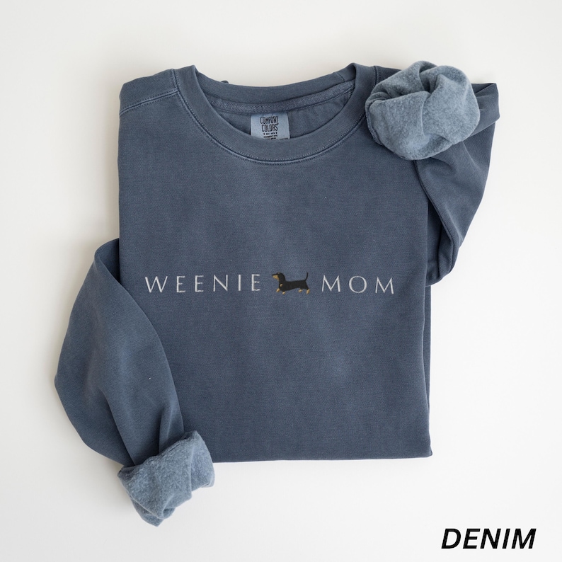 Puede incluir: Una sudadera azul denim con las palabras "WEENIE MOM" en letras blancas y una peque&ntilde;a ilustraci&oacute;n de perro salchicha. La sudadera tiene cuello redondo y mangas largas con pu&ntilde;os. La palabra "DENIM" est&aacute; impresa en la parte inferior.