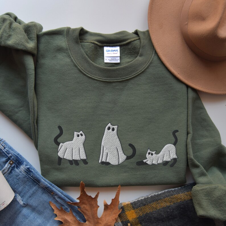 K&ouml;nnte beinhalten: Ein gr&uuml;nes Sweatshirt mit einem wei&szlig;en Geisterkatzen-Design. Das Design zeigt drei Katzen, die als Geister verkleidet sind. Das Sweatshirt liegt auf einem wei&szlig;en Hintergrund mit einem braunen Hut und einer blauen Jeans.
